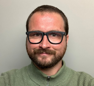 Man with short dark hair wearing black rimmed glasses and a green sweater.