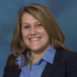Woman with dark blonde hair wearing a black blazer over a blue collared shirt.
