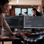 Two professionals working over a desktop computer that features code editing on the screen.