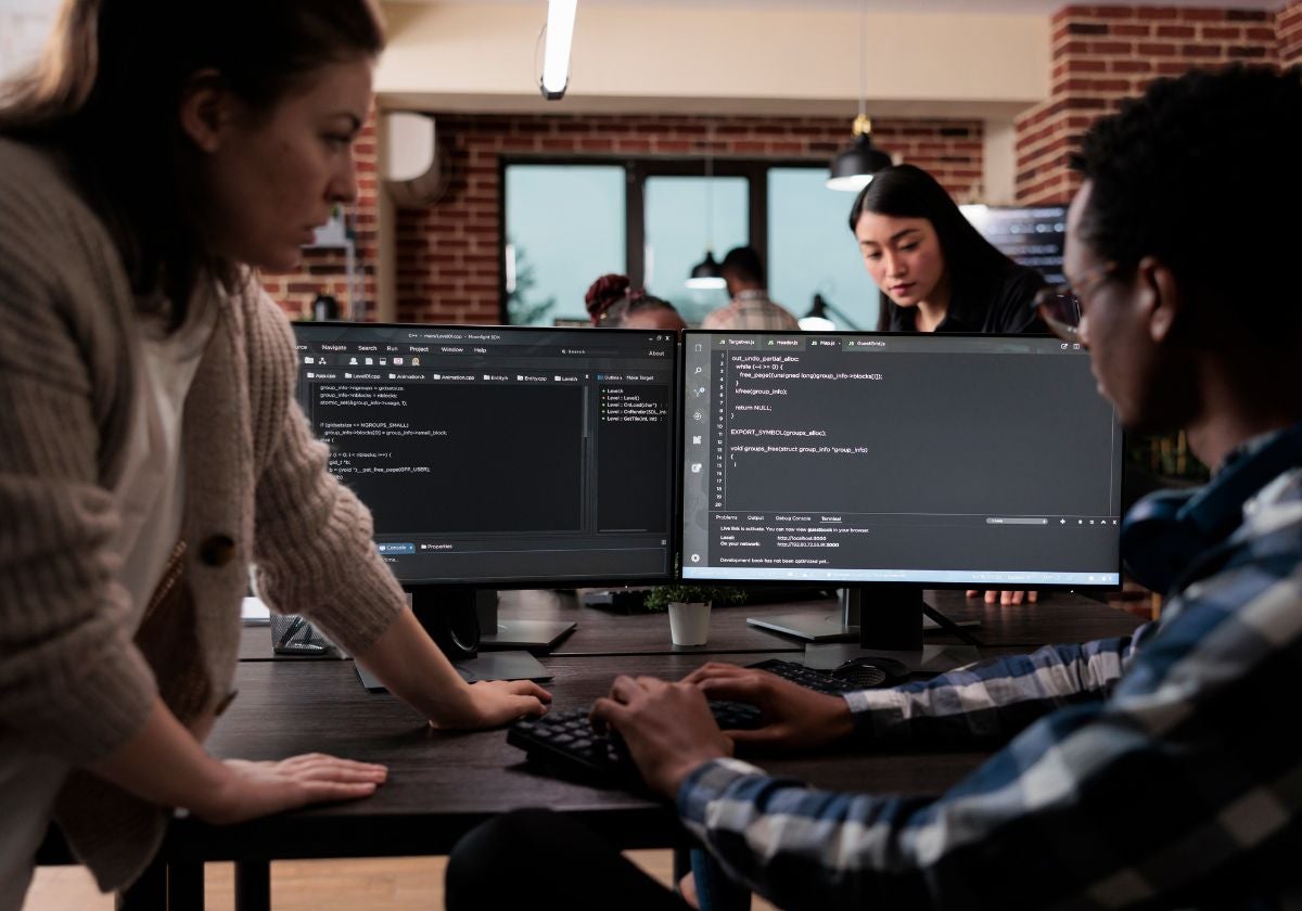 Two professionals working over a desktop computer that features code editing on the screen.