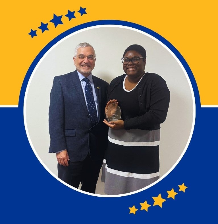 A man with gray hair and glasses poses with a woman with dark skin and glasses who holds a clear glass award in her hands