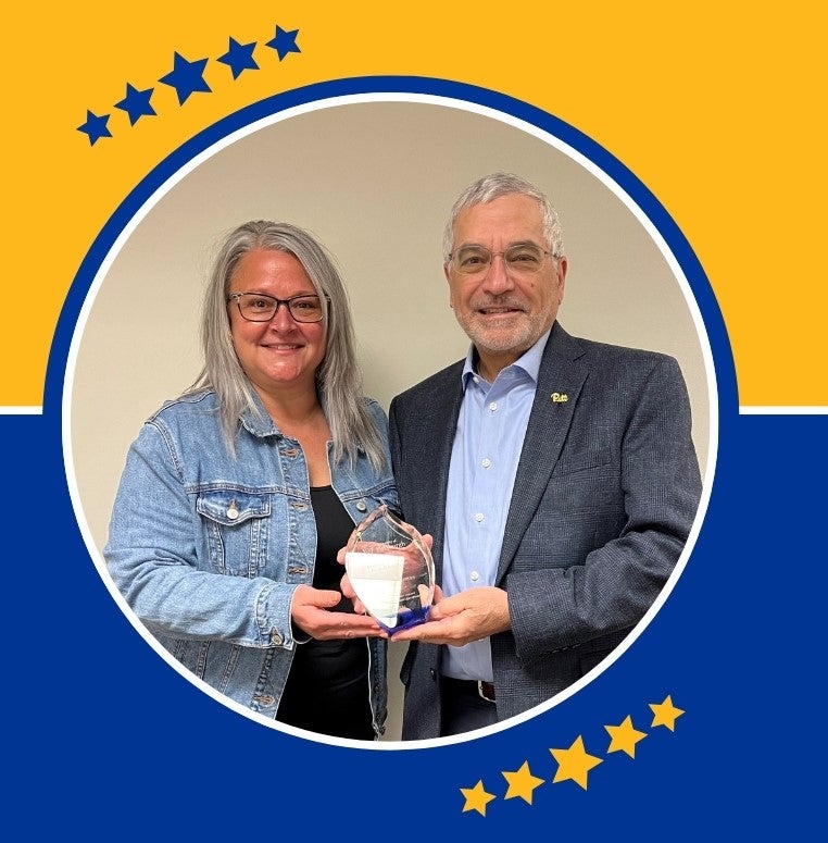 A woman with long gray hair and glasses poses with a man and gray hair and glasses as they both hold a clear glass award in their hands