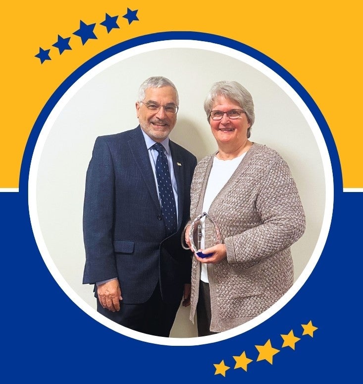 A man with gray hair and glasses poses with a woman with gray hair and glasses as she holds a clear glass award in her hands