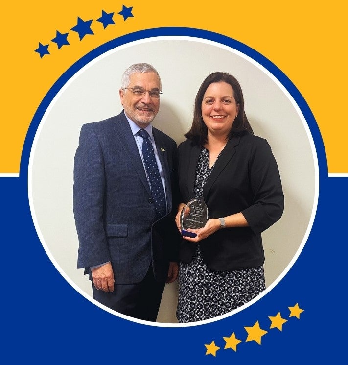 A man with gray hair and glasses poses with a woman with long dark hair holding a clear glass award in her hands