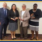 A man and three women holding clear glass awards pose for a group photo