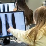 A young woman with long blonde hair and her back turned points at a computer monitor with her left hand pointing at radiograph images of a knee.