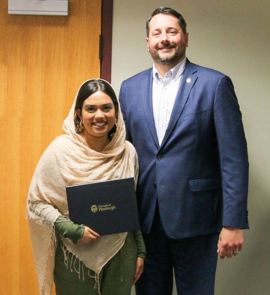 A young woman wearing a tan head scarf holds a certificate and stands with a tall man in a blue suit