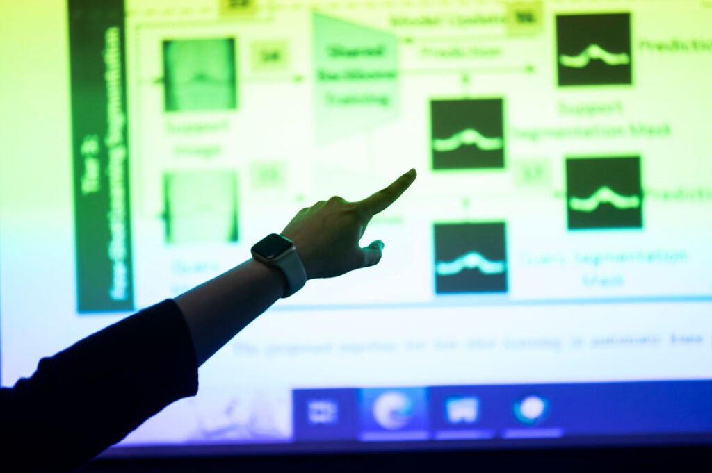 An arm and finger point to a projected screen showing blurred images of data