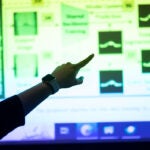 An arm and finger point to a projected screen showing blurred images of data
