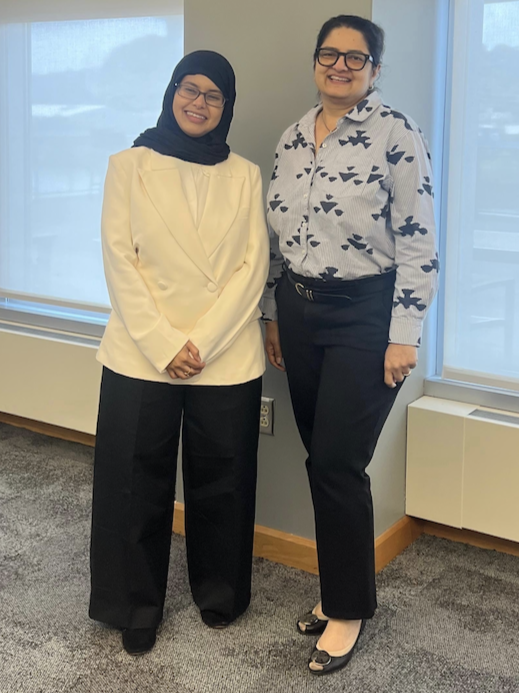 Photograph of two women standing indoors in front of large windows with blinds, smiling at the camera. One woman wears a white blazer with black pants and a black hijab, while the other wears a patterned white blouse with black pants and glasses.