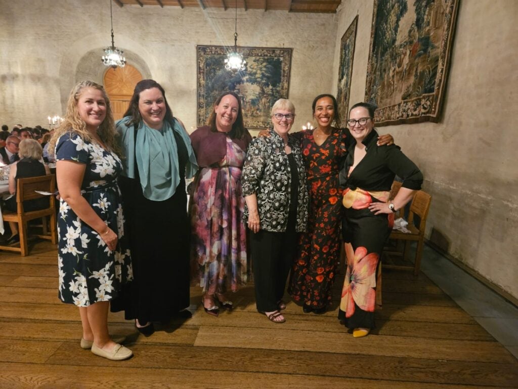 A group of six women in colorful dress stand in a large hall with tapestries on the walls