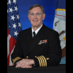 A man wears a formal black U.S. Navy coat with four gold bands on the sleeves and colorful military pins on the lapel.
