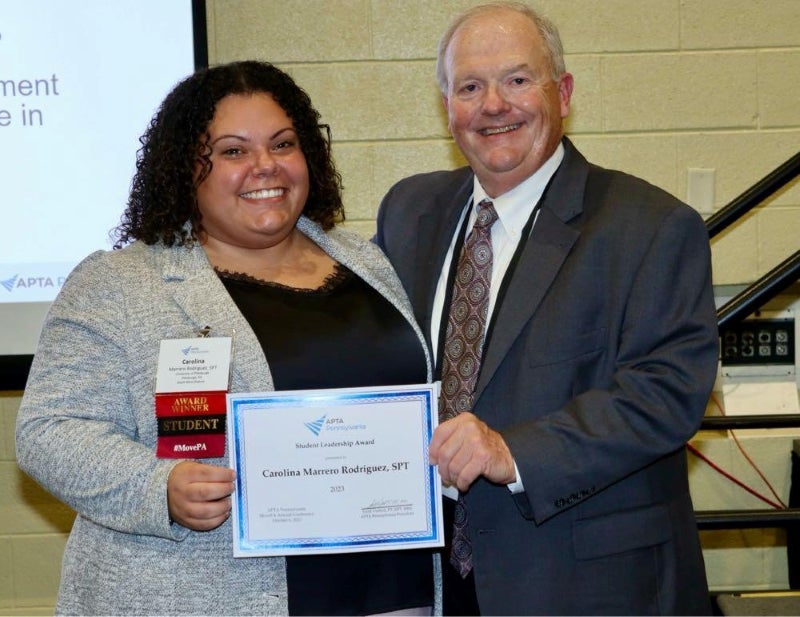 A woman with brown curly hair and a gray jacket smiles while holding a certificate with a man in a suit