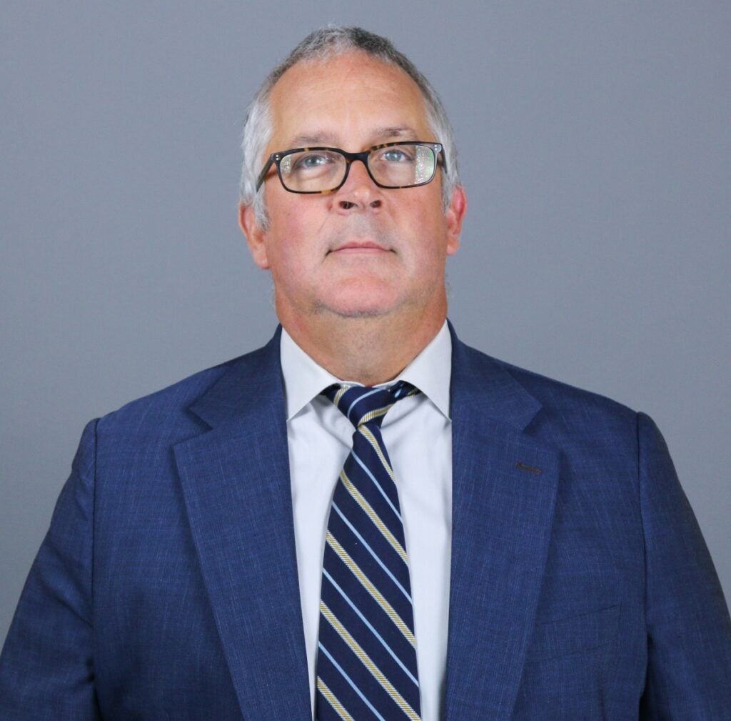 A man with short gray hair and glasses wears a blue dress jacket, white shirt and blue tie with yellow stripes