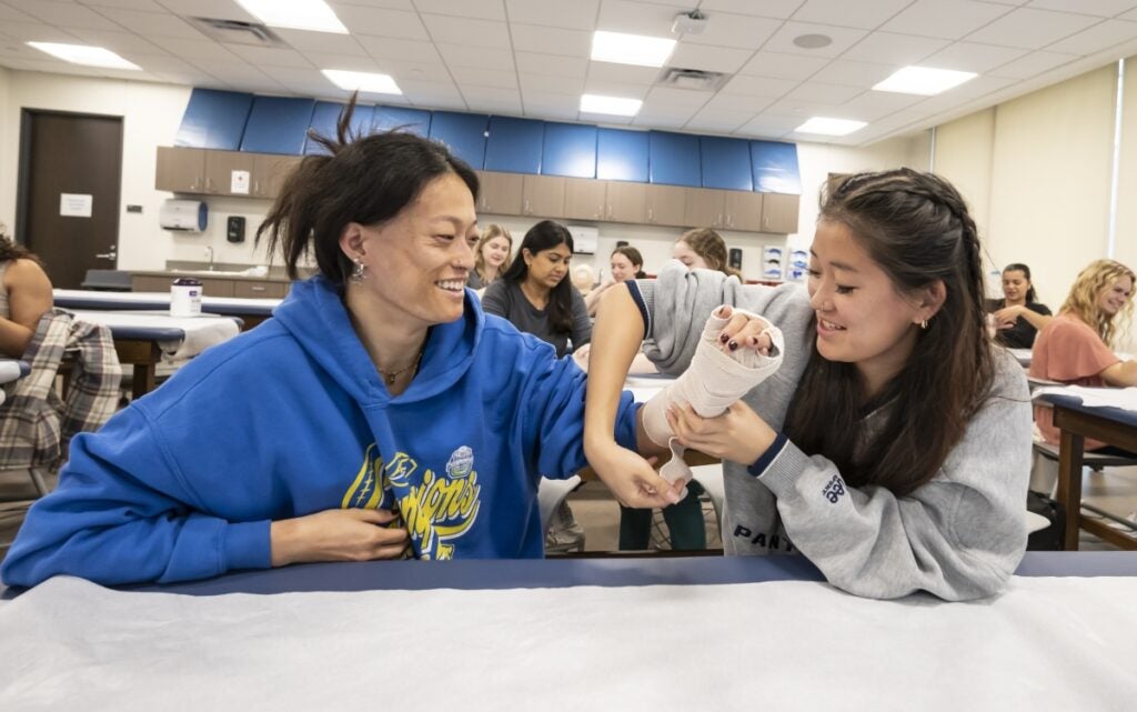 Two women sit at a table practicing wrapping an arm with a splint