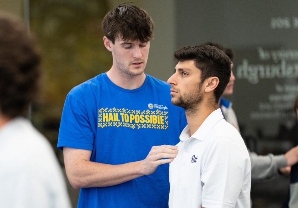 A man with dark brown hair wearing a blue t-shirt performing a chiropractic adjustment on a man with dark brown hair and facial hair wearing a white collared shirt.
