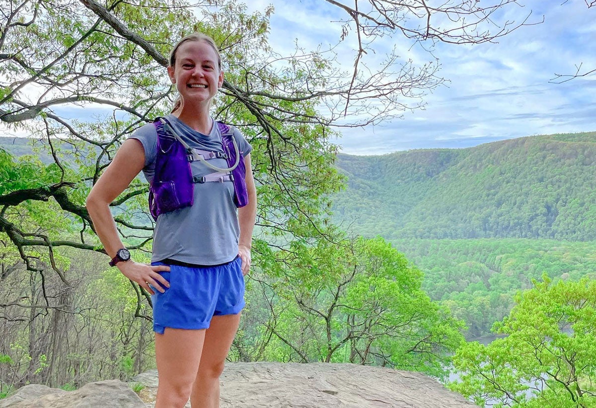 A woman wearing running clothing and a hydration vest smiles standing on a rocky overlook with a river and hills in the background