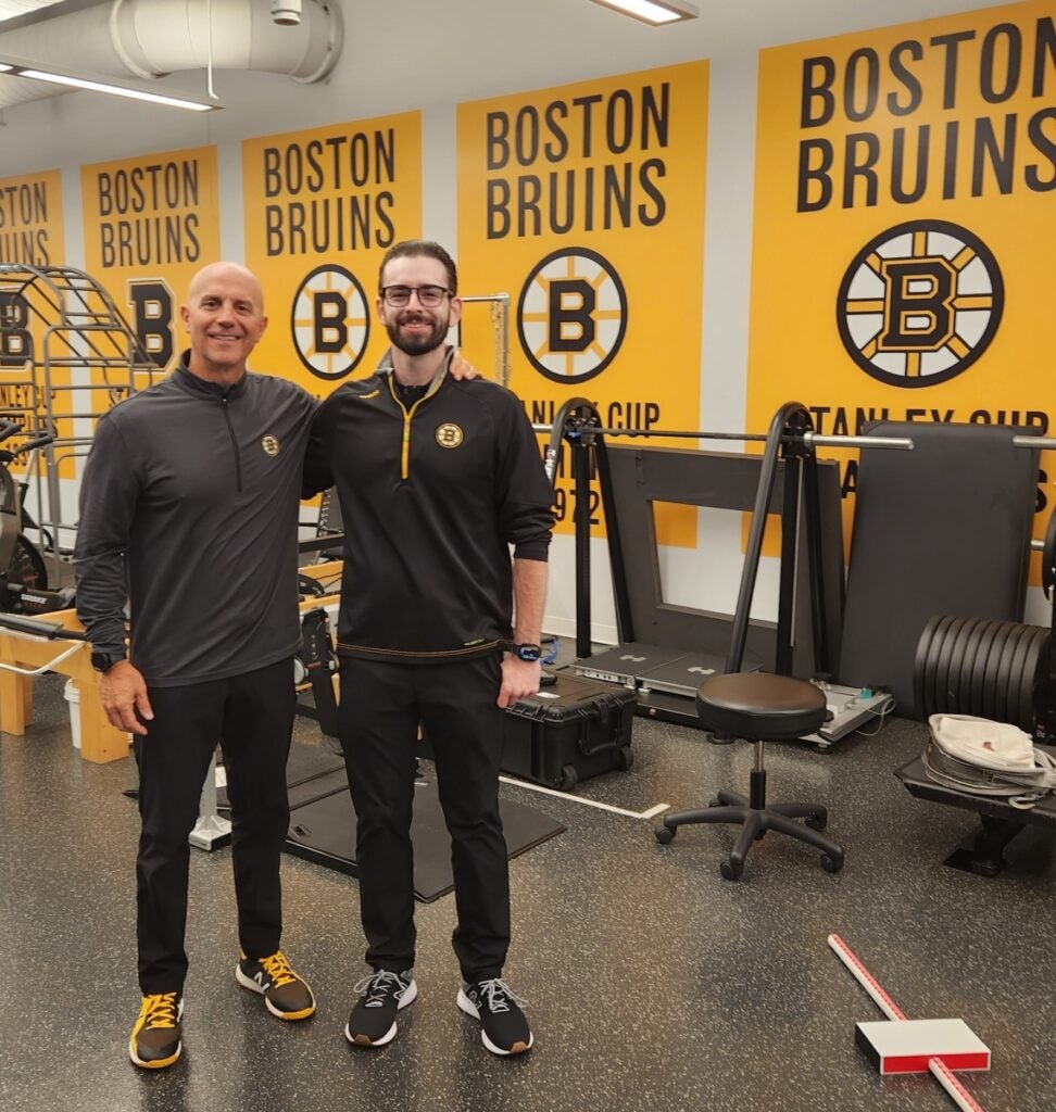 Two men wearing black and gray workout clothes post in front of workout equipment with Boston Bruins signs on the wall behind them