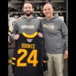 Two men wearing gray sweatshirts saying Bruins Hockey while the man with a beard hold a black hockey jersey with gold letters saying Koontz and the number 24