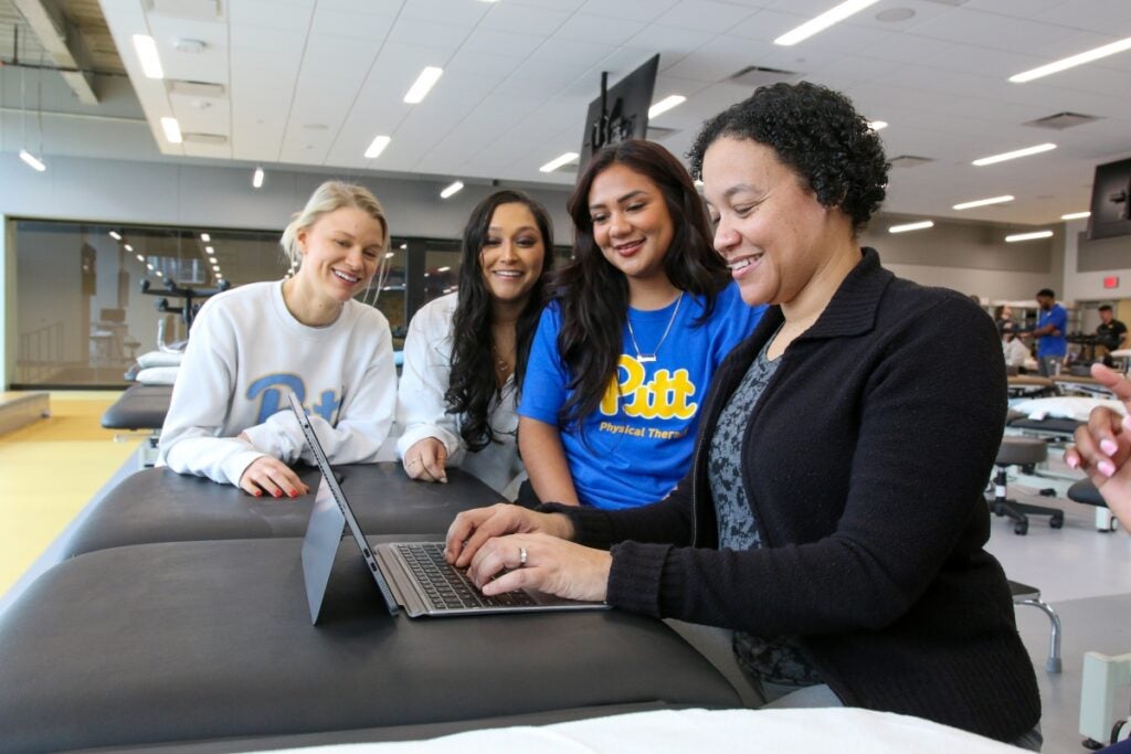 Three female students sit next to a female professor as she types on a laptop computer