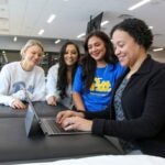 Three female students sit next to a female professor as she types on a laptop computer