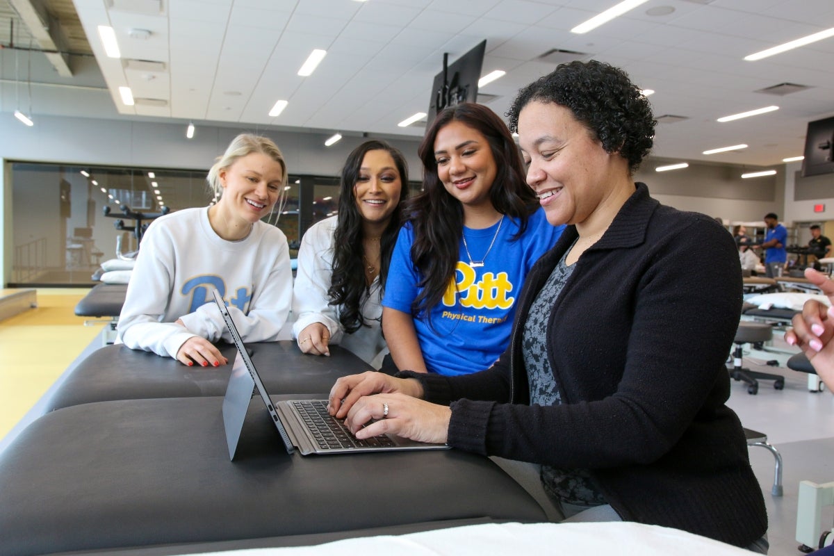 Three female students sit next to a female professor as she types on a laptop computer