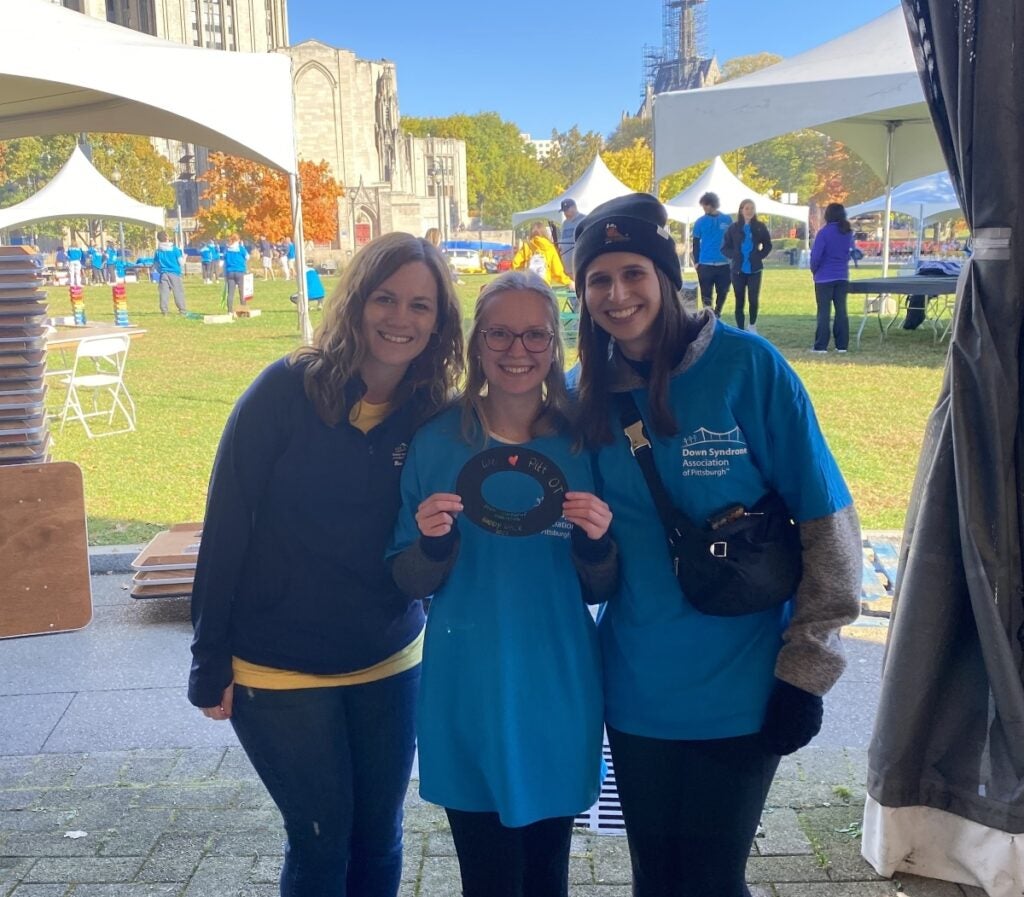 Three women face the camera smiling with the Cathedral of Learning and lawn in the background