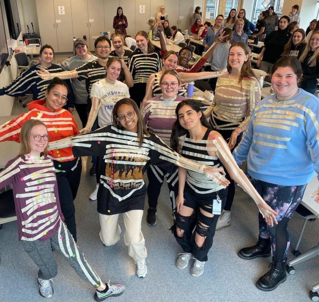 A group of students stands facing the camera, they all have strips of masking tape down their arms and across their torsos
