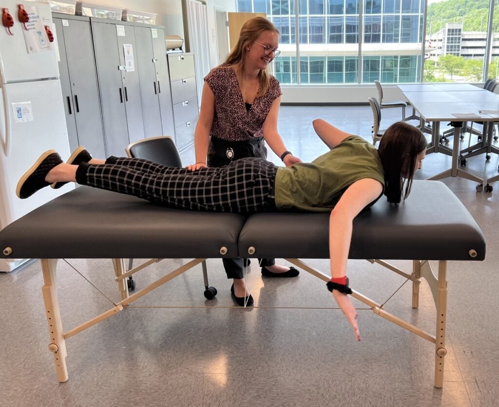 A woman with blonde hair stands next to a therapy table while another woman lays on the table moving her legs and arms