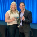 A woman with long blonde hair wearing a sleeveless cream top holds a large silver trophy cup with a man with short dark hair wearing a dark navy suit