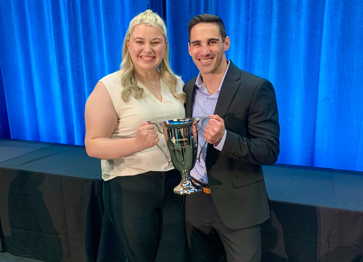 A woman with long blonde hair wearing a sleeveless cream top holds a large silver trophy cup with a man with short dark hair wearing a dark navy suit