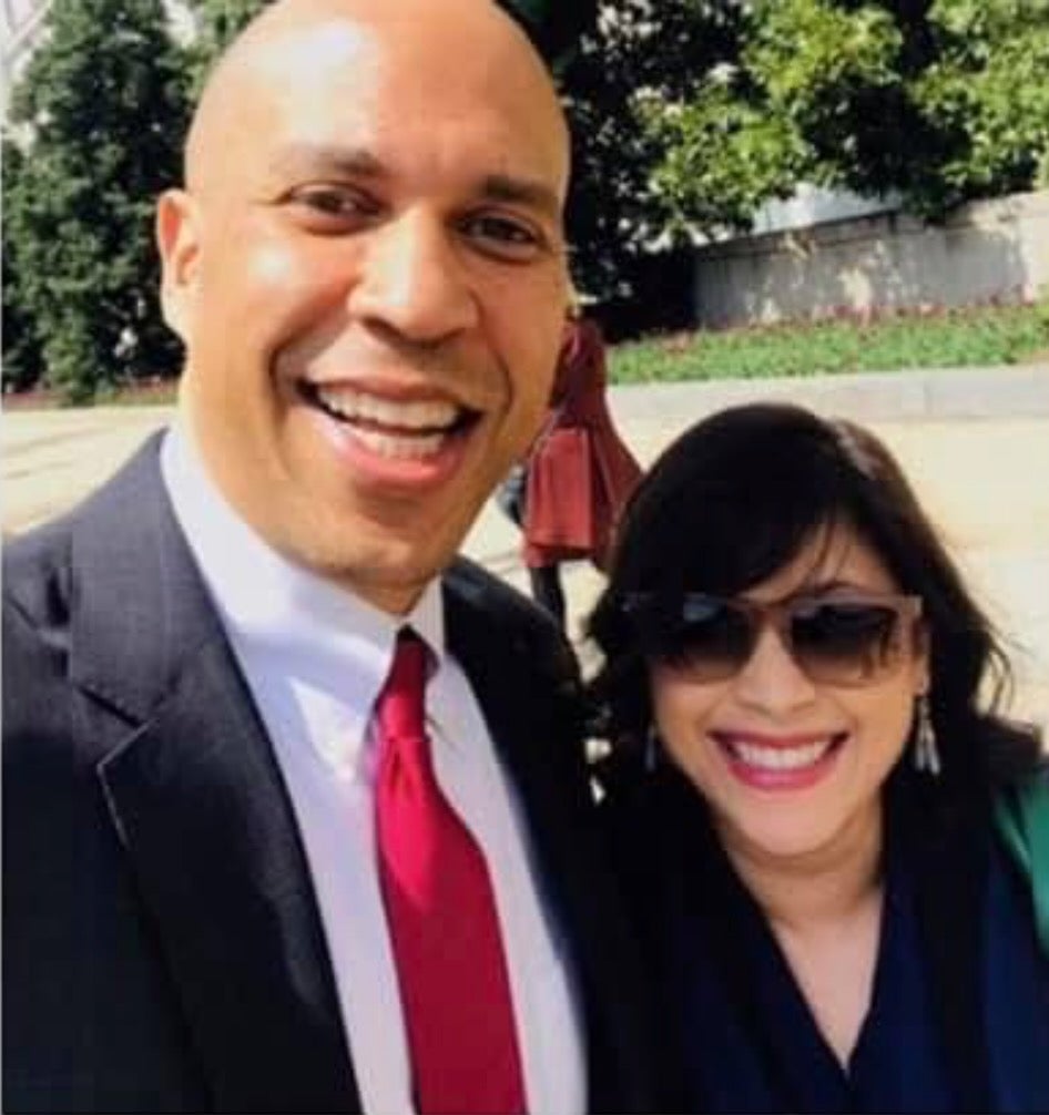 A bald man wearing a dark gray suit jacket over a white collared shirt and a red tie standing next to a woman with short dark brown hair wearing a navy blue shirt and brown sunglasses.