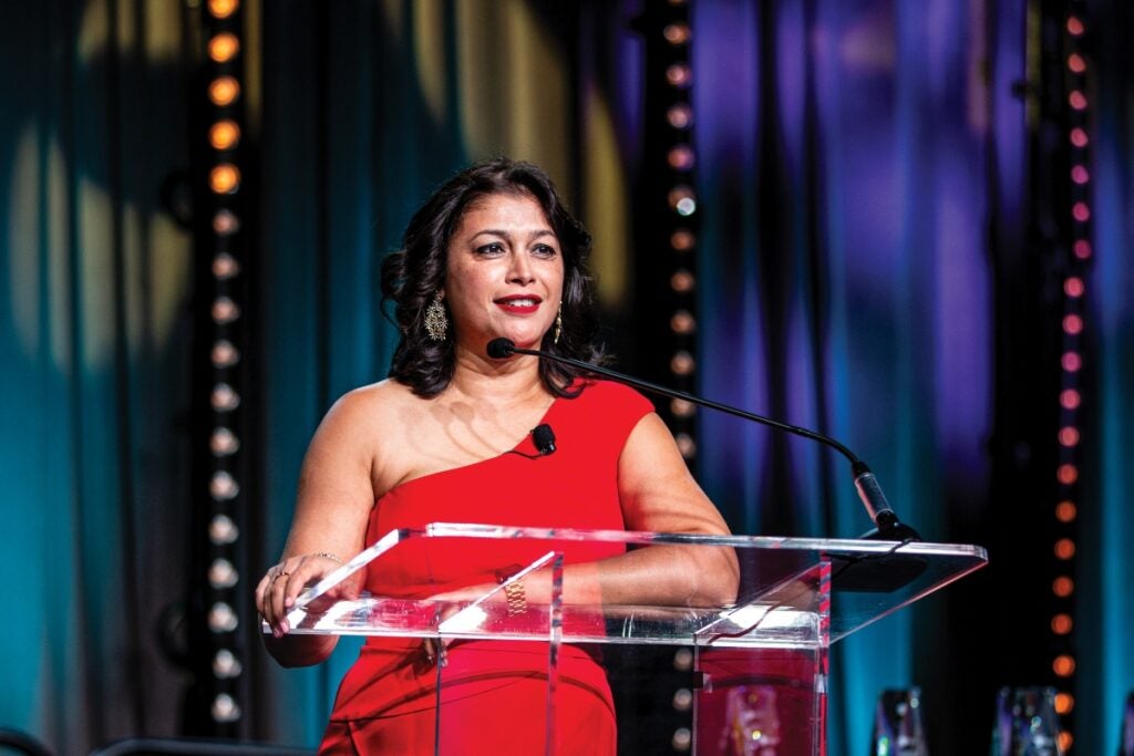 A woman with medium length brown hair wearing a red dress stands at a podium on a stage