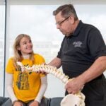 A female student with long blonde hair and a bright yellow t-shirts smiles up at a professor with gray hair who holds a model of a spine
