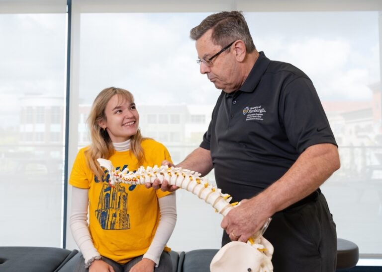 A female student with long blonde hair and a bright yellow t-shirts smiles up at a professor with gray hair who holds a model of a spine