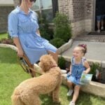A woman in a blue dress and sunglasses smiles at a young girl sitting on a step in front of a curly haired dog