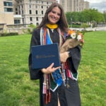 A woman with long brown hair wears graduation regalia and hold a diploma