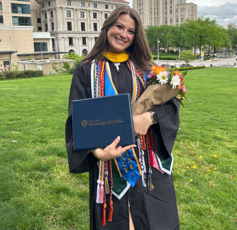 A woman with long brown hair wears graduation regalia and hold a diploma