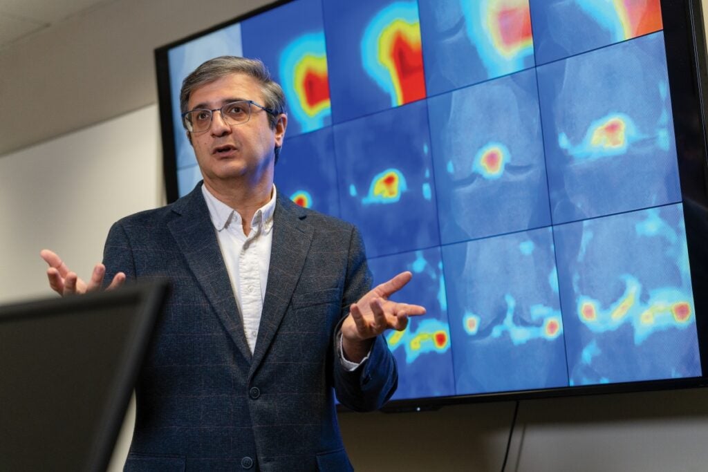 A man wearing a jacket and glasses speaks in front of a digital monitor showing shapes in different shades of blue, yellow and red