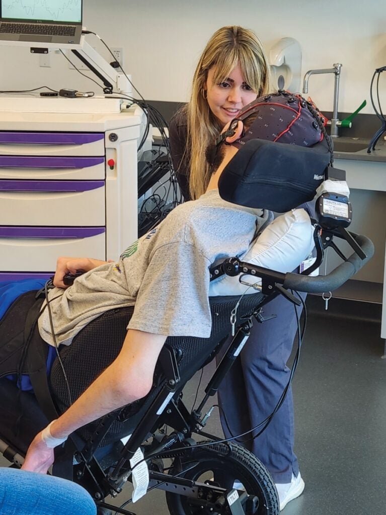 A woman with long blond hair talks to a man in a wheelchair wearing a cap on his head with electronic sensors.