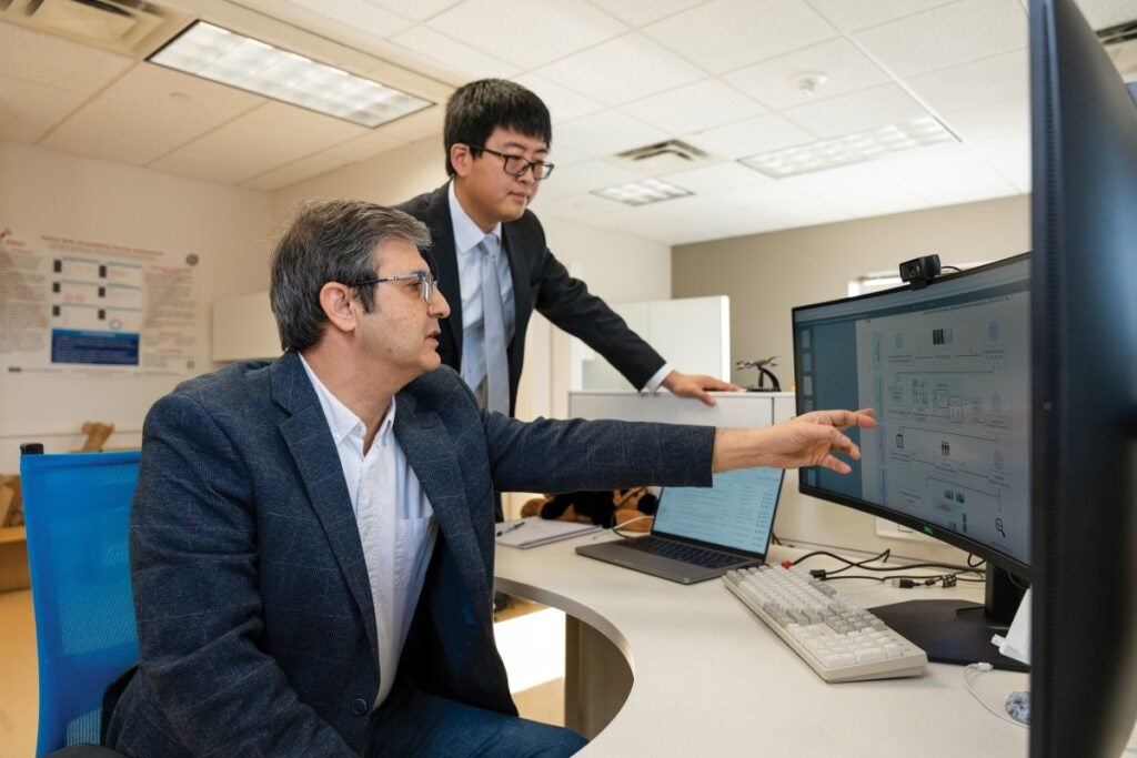 One man sits at a computer desk pointing to a monitor and talking while another man stands next to him