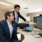One man sits at a computer desk pointing to a monitor and talking while another man stands next to him