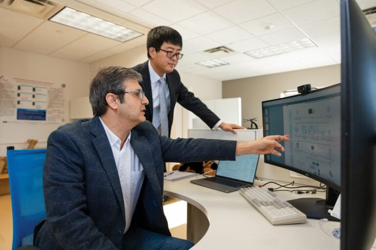 One man sits at a computer desk pointing to a monitor and talking while another man stands next to him