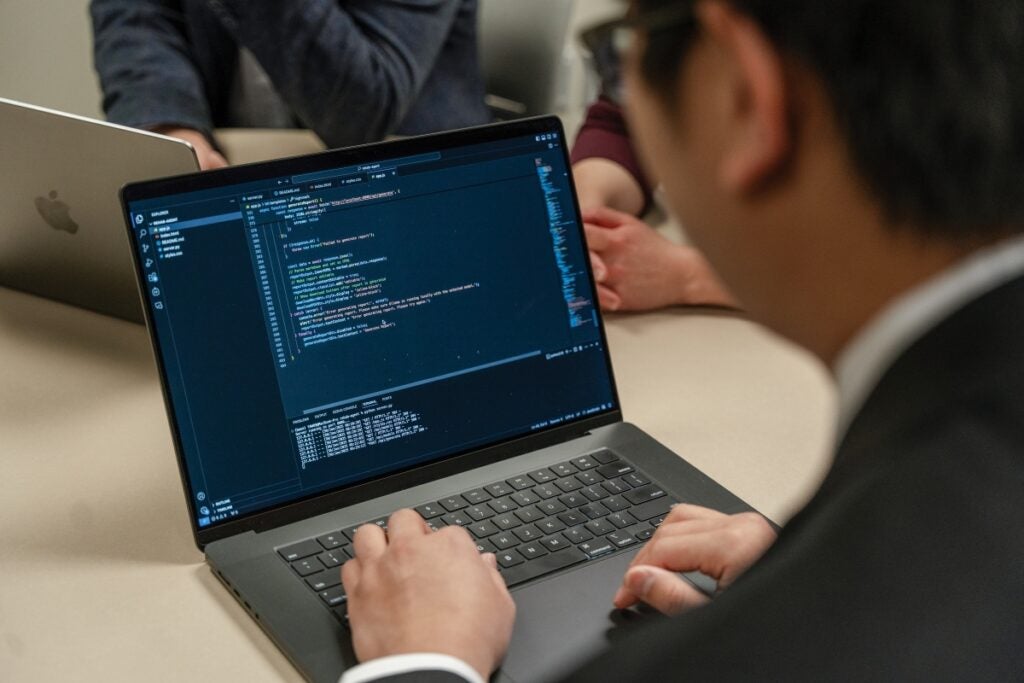 A man sits at a laptop computer that shows a screen of programming code.