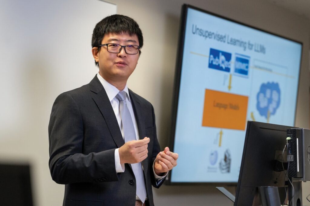 An Asian man with glasses wearing a suit and tie gives a lecture in front of a digital screen