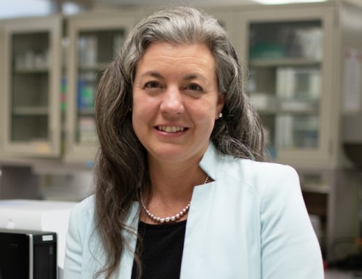 A woman with long gray hair wears a white jacket over a dark shirt and pearl necklace