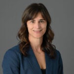 A portrait photo of a woman with long brown hair and short banks wearing a blue dress jacket