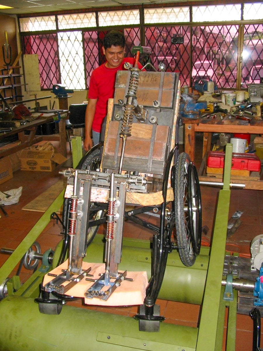 A man with medium skin tone wearing a red shirt stands in a workshop behind a large wheelchair with test equipment set up on the chair