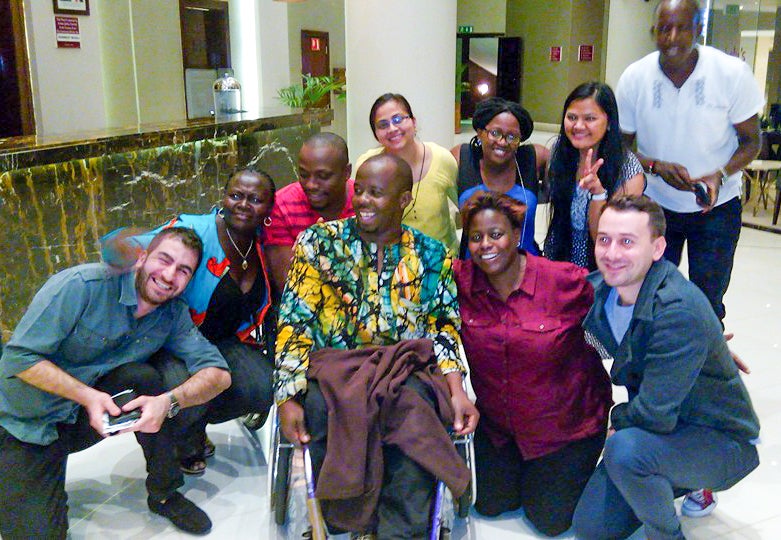A group of men and women with light and dark skin and colorful clothing surround a person in a wheelchair