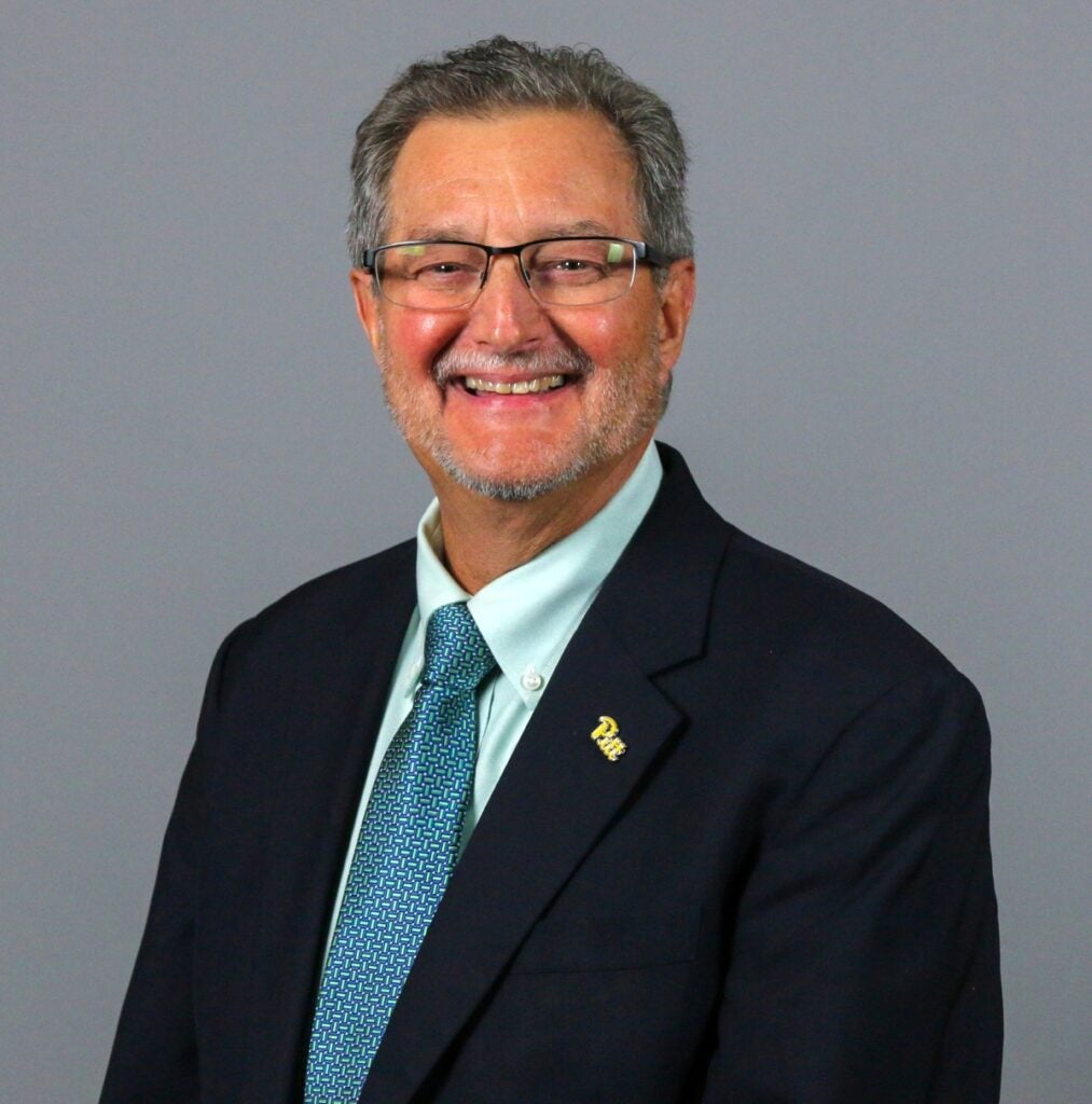 A man with short gray hair and glasses wears a dark navy jacket over a white dress shirt and blue checkered tie